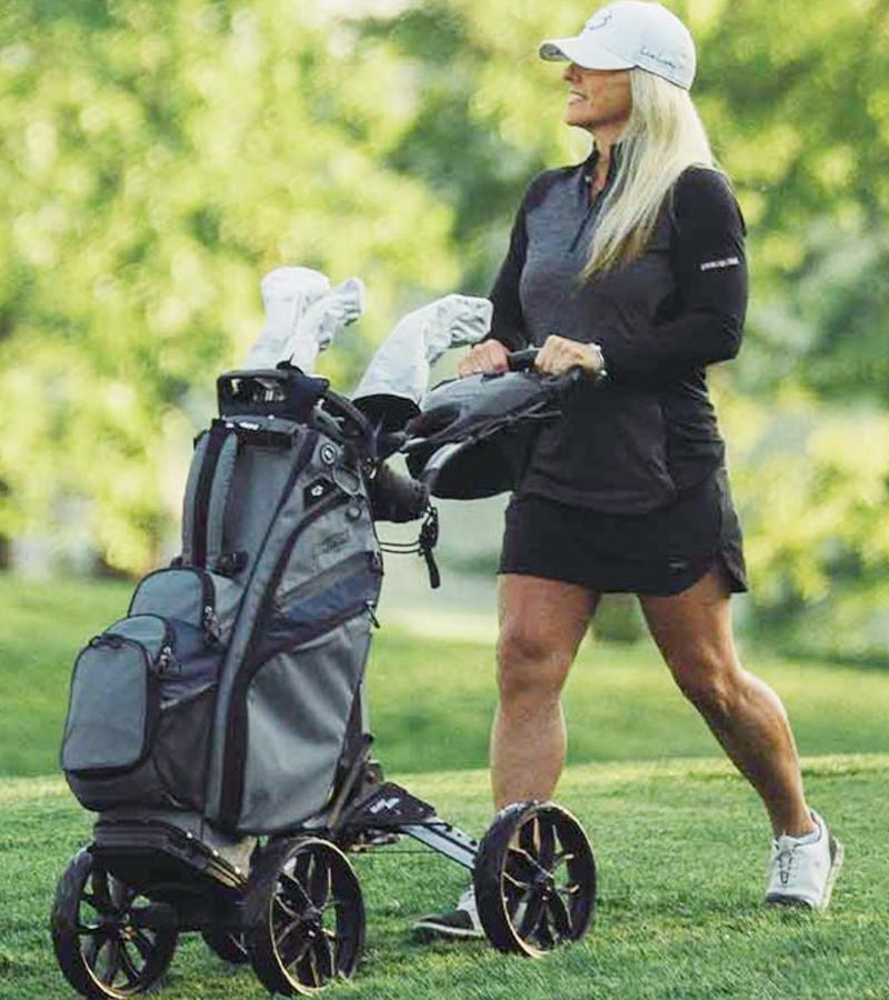 BagBoy Quad Pro Golf Push Cart – in Black/Slate - female golfer pushing cart with golf bag & clubs along a golf course