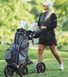 BagBoy Quad Pro Golf Push Cart – in Black/Slate - female golfer pushing cart with golf bag & clubs along a golf course