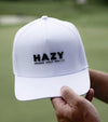 Hazy Golf: Hazy X Tremont Dune Hat  in white front view with logo across the front crown in black. Hat held by hands of golfer against an out of focus green background