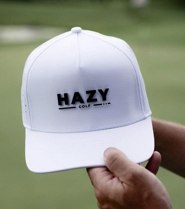 Hazy Golf: Hazy X Tremont Dune Hat  in white front view with logo across the front crown in black. Hat held by hands of golfer against an out of focus green background